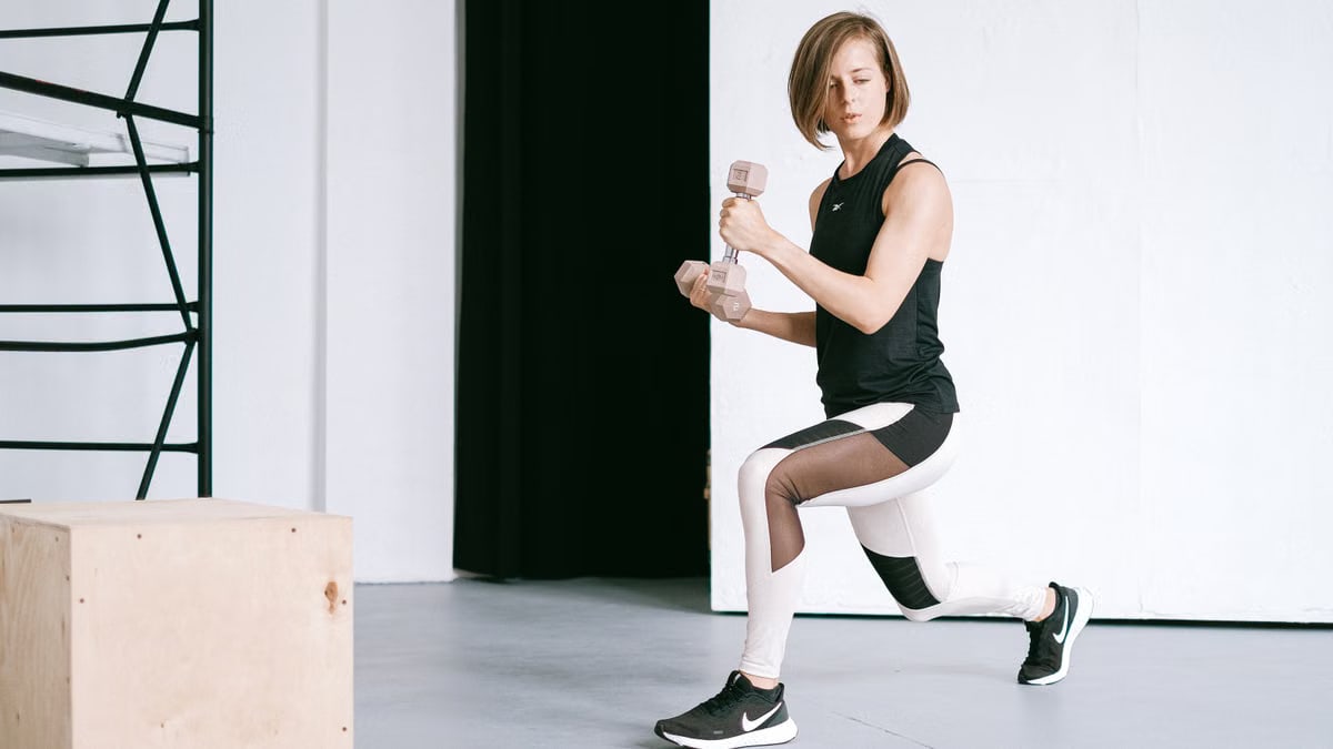 A young woman performing lunges with dumbbells in a modern indoor gym setting.
